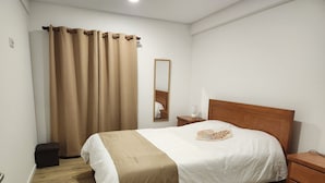A neatly made bed with a wooden headboard, a bedside table with a lamp, a mirror, and a brown curtain.