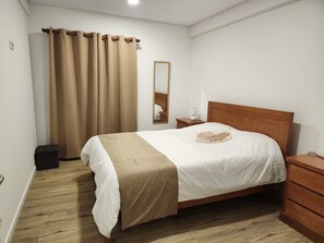 Un lit bien fait, avec une tête de lit en bois, une table de chevet avec une lampe, un miroir et un rideau marron.