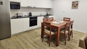 A modern kitchen with white cabinets, a stainless steel refrigerator, a microwave, a kettle, and a dining table with wooden chairs.