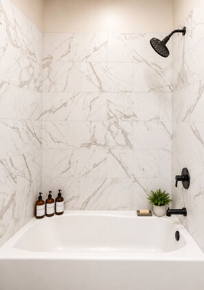 Une salle de bain avec un mur en marbre blanc, une pomme de douche noire, une baignoire blanche et trois bouteilles brunes de shampoing et d’après-shampoing.