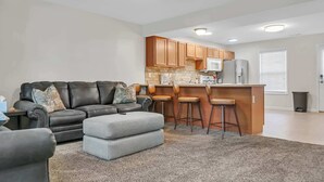 A modern living room with a sofa, ottoman, and bar stools, a kitchen with wooden cabinets and appliances, and a window with blinds. - Cozy Bloomington Townhouse Close to IU Campus (Bloomington)