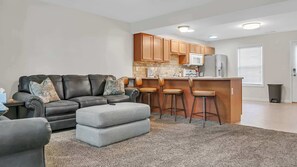 A modern living room with a sofa, ottoman, and bar stools, a kitchen with wooden cabinets and appliances, and a window with blinds. - Cozy Bloomington Townhouse Close to IU Campus (Bloomington)