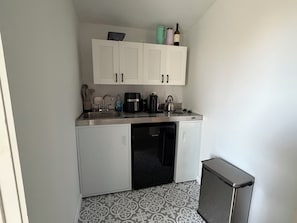 Una cocina compacta con armarios blancos, un lavamanos de acero inoxidable, una cafetera y un hervidor.