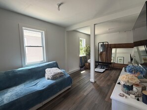 Una sala de estar con sofá azul, piso de madera, un espejo grande y una ventana con persianas.