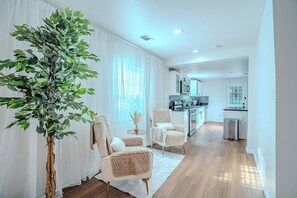 Ein modernes Wohnzimmer mit einer großen Pflanze, zwei Sessel, einem kleinen Tisch und einer Küche im Hintergrund.