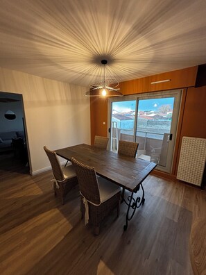 Comedor con mesa de madera y sillas de mimbre, lámpara de techo y vista al exterior por una puerta de vidrio.