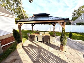 En overdækket terrasse med et bord og stole, omgivet af et hvidt hegn og grønne planter.