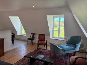Ein Zimmer mit schrägem Dach, zwei Fenstern, Holzboden, einem dunkelbraunen Tisch und einem blauen Stuhl.