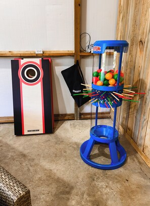 Une machine de flipper bleue, avec un distributeur de billes rempli de billes et de quilles colorées, un sac noir sur lequel on peut lire « BACKYARD HERO », et une machine de flipper blanche et rouge en arriÚre-plan.