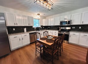 A kitchen with white cabinets, a wooden dining table with chairs, a chandelier, and a window with blinds.