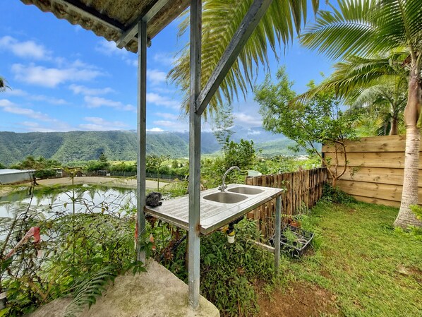 Un espacio al aire libre cubierto con una terraza de madera, un lavamanos de metal y vistas a un lago y montañas.