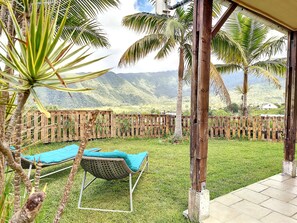 A covered outdoor area with a wicker lounge chair, a wooden fence, and palm trees. - Le Yucca - Terrasse - TV (Saint-Louis)