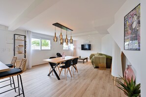 Een moderne woonkamer met een eethoek, een aan de muur bevestigde televisie en een groot raam.