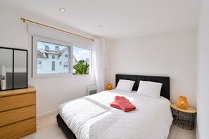 Een moderne slaapkamer met een groot bed, een houten dressoir, een raam met uitzicht op gebouwen en een nachtkastje met een lamp.