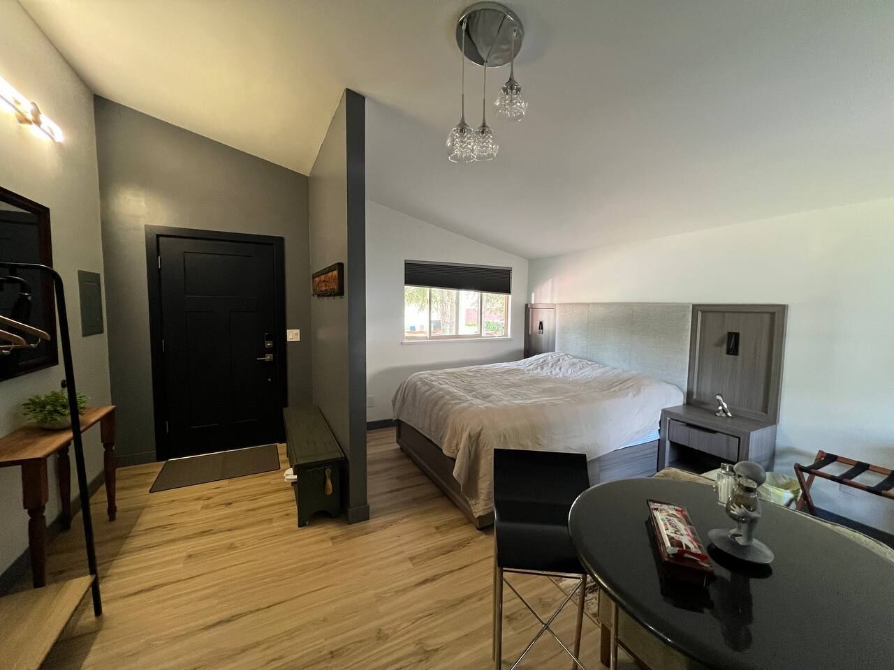 A modern hotel room with a bed, a desk, a chair, a small table, a desk lamp, a nightstand, a TV, and a window with blinds.