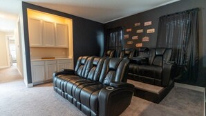 Un cine en casa con sofá y dos sillones reclinables, un área de cocina con armarios blancos y una pared decorada con placas.