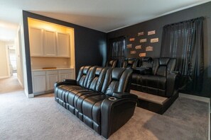 Ein Heimkino mit einer Couch und zwei Relaxsesseln, ein Küchenbereich mit weißen Schränken und eine Wand mit dekorativen Plaketten.