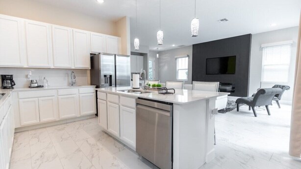 Una cocina moderna con armarios blancos, una isla central, electrodomésticos de acero inoxidable, iluminación colgante y un televisor de pantalla plana montado en la pared.