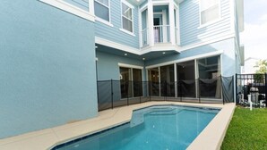 Una piscina con un porche techado, una terraza cercada y una casa con fachada azul y ventanas amplias.
