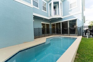 Ein Pool mit überdachter Terrasse, einem umzäunten Deck und einem Haus mit blauer Fassade und großen Fenstern.