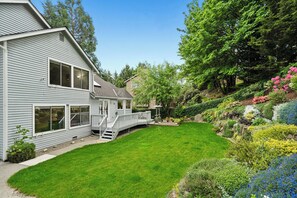 Ett tvåvåningshus med en inglasad veranda, en välskött gräsmatta och en mängd olika blommande växter.