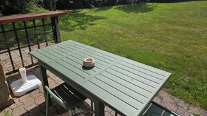 Et grønt metalbord med en hvid skål, placeret på en belagt plads med græs og træer i baggrunden.