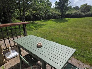 Una mesa metálica verde con un tazón blanco encima, ubicada en una zona pavimentada con césped y árboles al fondo.