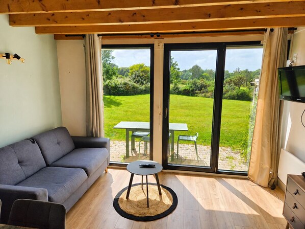 Habitación con sofá, una mesita con un control remoto, televisión y una puerta corrediza de vidrio a un jardín.