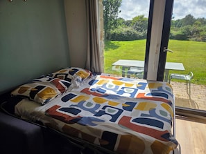 Una cama con un edredón de estampado geométrico colorido, una ventana que da a un área verde y una mesa afuera.
