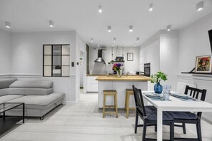 Eine moderne Küche mit einer zentralen Kochinsel, ein Essbereich mit weißem Tisch und Stühlen sowie ein graues Sofa in einem großzügigen Wohnbereich.