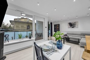 A modern living room with a dining area, a sofa, and a view of a building through a large window. - Wola Apt - Parking, Balcony, Workspace - by Rentujemy (Warszawa)