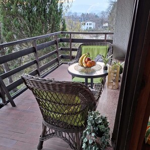 Un balcon avec des fauteuils en osier, une petite table ronde sur laquelle est posé un panier de fruits, et une vue sur les arbres et les bâtiments.