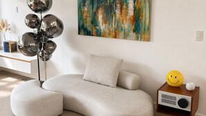 Een moderne woonkamer met een hoekbank, een rond poefje, een vloerlamp met reflecterende bolletjes, een schilderij aan de muur en een klein bijzettafeltje met een speaker en een decoratief object.