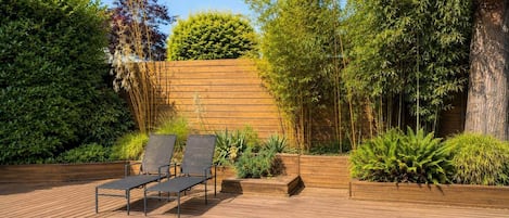 A wooden deck with two lounge chairs, surrounded by lush greenery and a wooden wall.