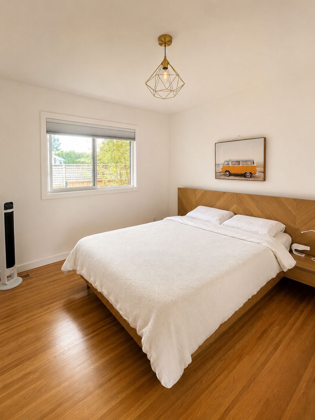 A bedroom with a wooden bed, a bedside table, a wall-mounted light fixture, a window with blinds, and a framed picture on the wall.