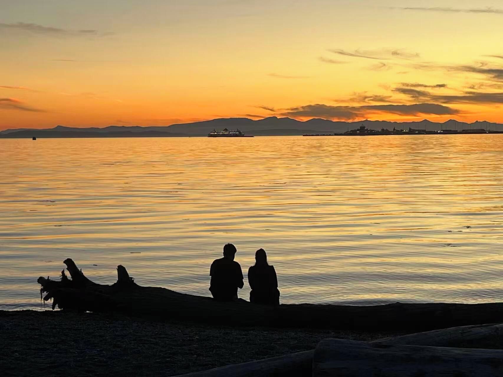 Ein Sonnenuntergang über ruhigem Wasser, im Hintergrund sind Silhouetten von Personen und ein Schiff zu sehen.