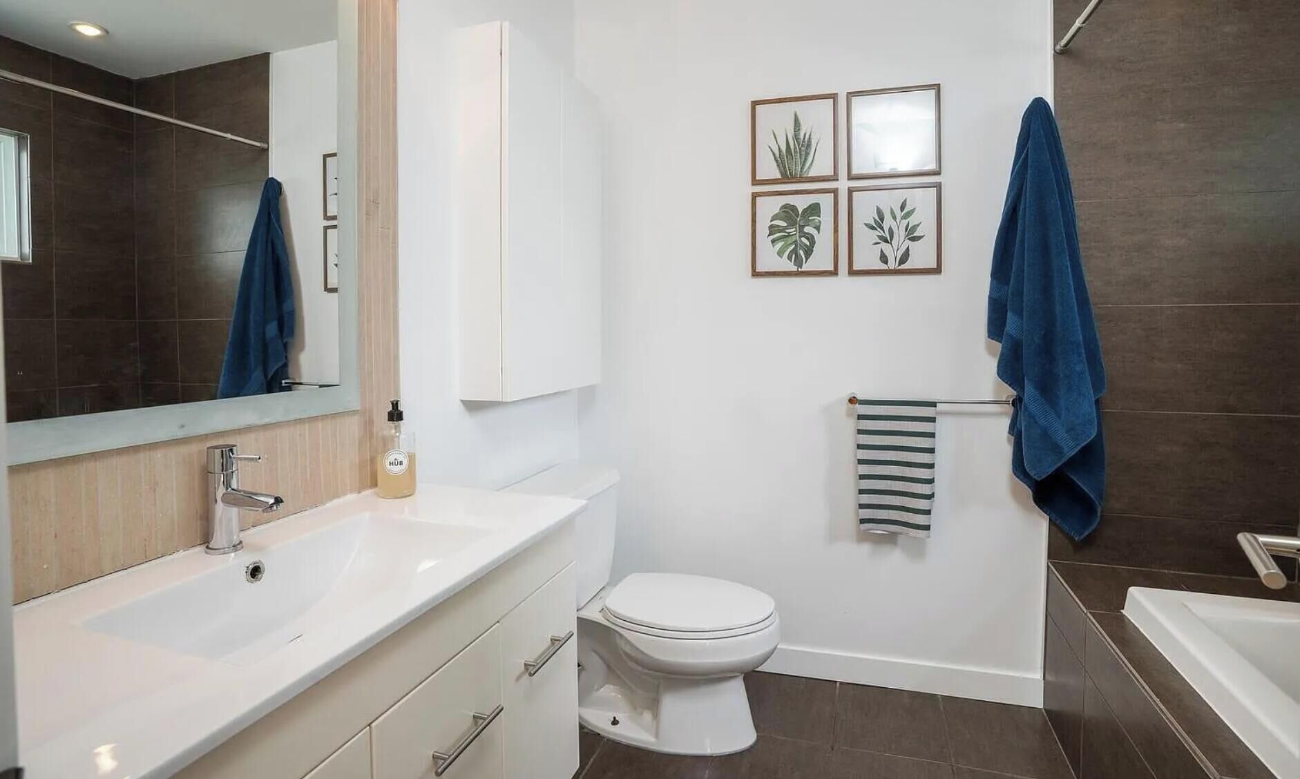 Een moderne badkamer met een wit wastafelmeubel, toilet en een badkuip. Op de muur hangen afbeeldingen van planten in lijsten en een blauwe handdoek aan de handdoekhouder.