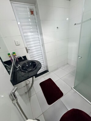 Un baño con un lavabo negro, paredes de azulejos blancos, cabina de ducha de cristal y una alfombrilla de baño roja.