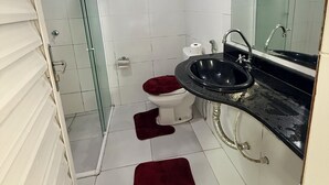 A compact bathroom with a glass shower enclosure, a white toilet, a black sink, and a red bath mat. - Rooms for rent in Belém (Belém)