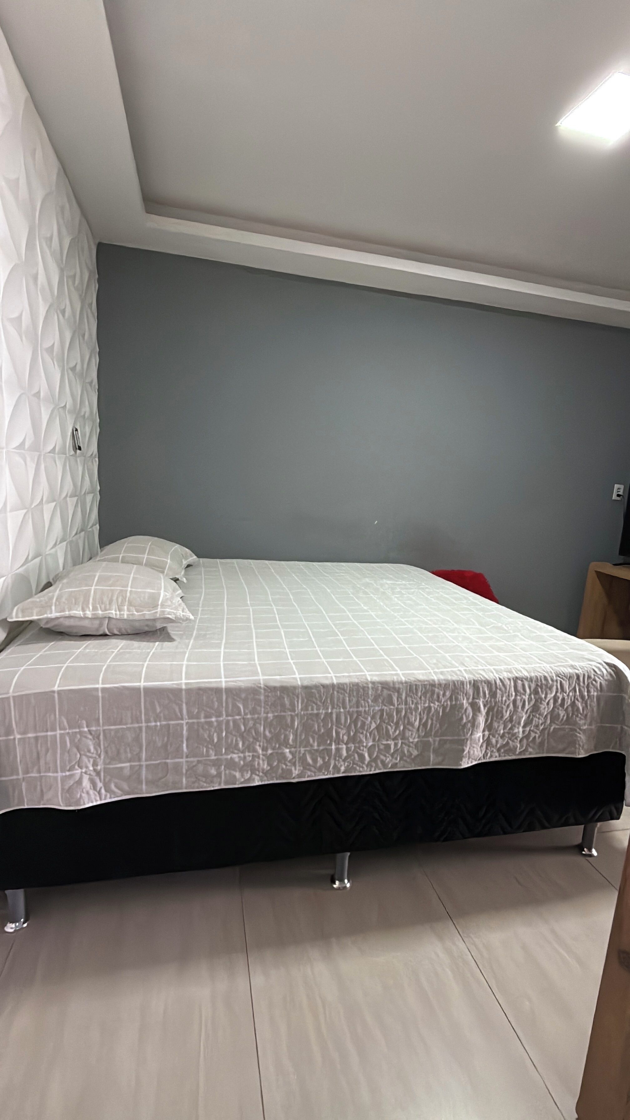 Un dormitorio con cama, una silla roja, una mesita de noche de madera y una lámpara de pared.