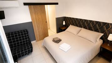 A hotel room with a bed, a nightstand, a lamp, a heater, and a window with curtains.