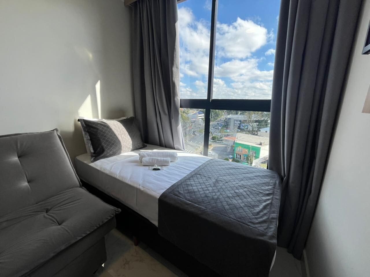 Quarto com uma cama, um sofá cinza e janela com vista para a cidade.