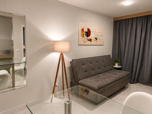 A modern living room with a glass coffee table, a grey sofa, a floor lamp, a painting, and a grey curtain.