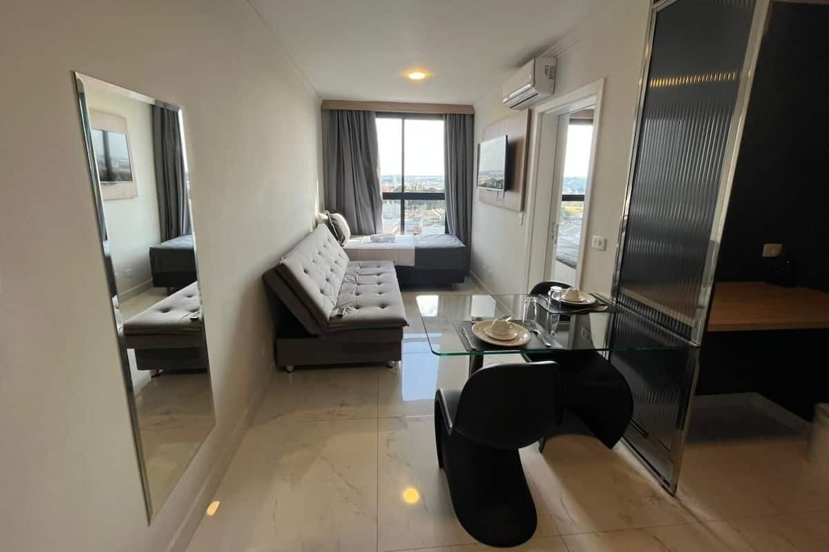 A compact hotel room with a sofa, a glass dining table, a TV mounted on the wall, and a large window offering a city view.
