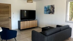 Un salón moderno con una televisión de pantalla plana, un sofá oscuro, un sillón azul, un mueble de madera y un ventanal grande.