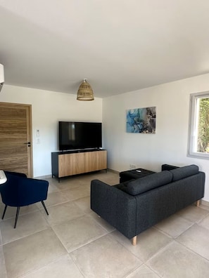 A modern living room with a flat-screen TV, a dark sofa, a blue chair, a wooden cabinet, and a large window. - LE SOLEIL DU GOLF PMR Sud (Saumane-de-Vaucluse)