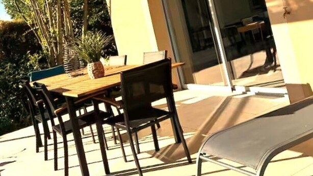 Un patio al aire libre con una mesa y sillas de madera, una sombra tejida y una puerta de cristal que da a un espacio interior.