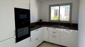 Una cocina con armarios blancos, horno negro, ventana con vistas y suelo de baldosas.