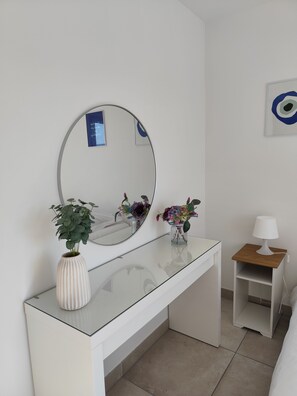 Ett vitt rum med en rund spegel, ett skrivbord med glasskiva, ett litet träbord som används som nattduksbord med en lampa, och en vas med blommor.