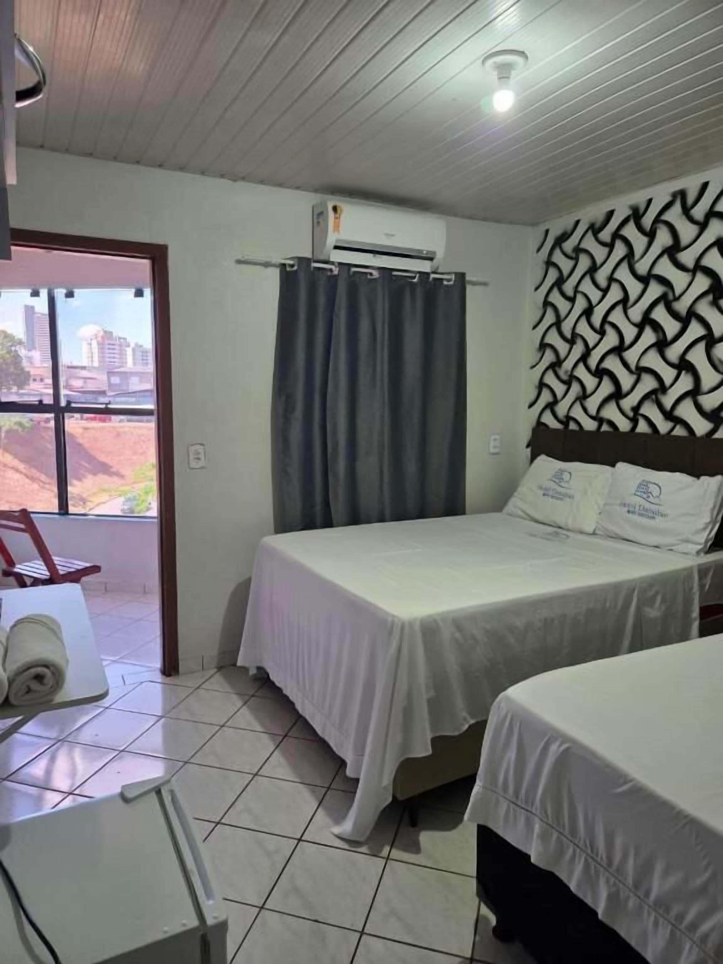 A hotel room with two beds, a window with curtains, an air conditioning unit, and a view of buildings outside.